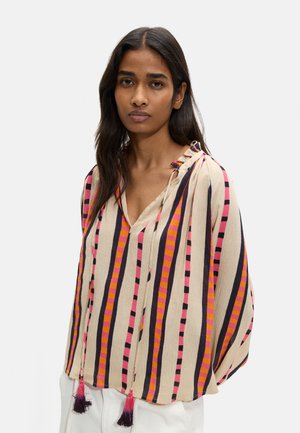 Kobieta o długich ciemnych włosach, ubrana w luźną bluzkę w pionowe paski w kolorach różowym, pomarańczowym, czarnym i beżowym oraz białe spodnie.