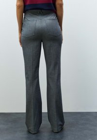 Personne portant un pantalon gris sur mesure et un haut bordeaux, debout sur un sol sombre devant un mur clair uni, vue de dos.