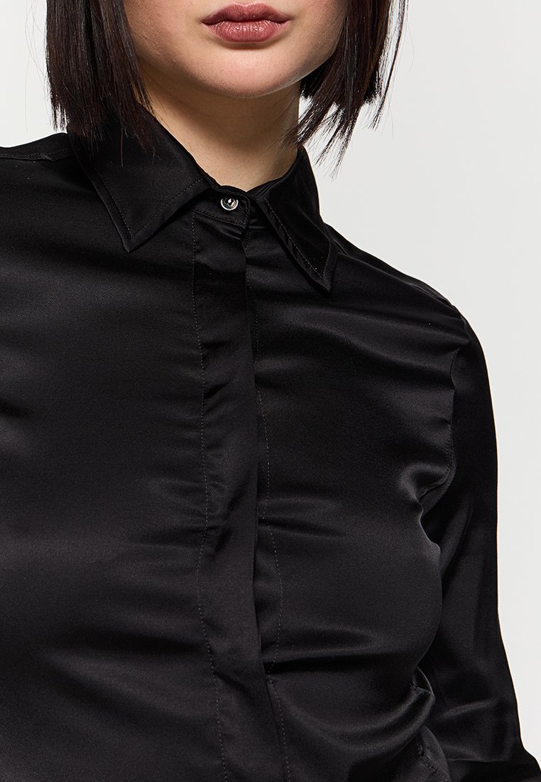 Schwarze Satinbluse mit klassischem Kragen und Knopfverschluss, mit glatter Oberfläche und dezentem Glanz. Kurzärmlig. Ohne Muster oder Akzente.
