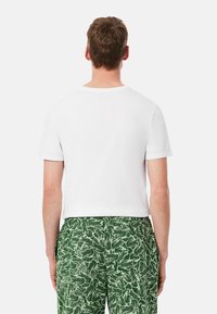 T-shirt bianca a maniche corte con collo rotondo, abbinata a pantaloni verdi con un motivo di piante stilizzate. Vista da dietro.