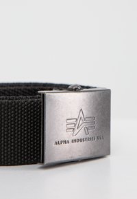 Cintura de nylon preta com uma fivela de metal prateada que apresenta um logótipo em relevo, formato retangular e um design texturizado em tecido.