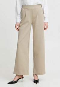 Beige vida byxor med hög elastisk midjeband, med frontveck. Bärs med svarta spetsiga klackskor. Slät, lättviktsmaterial.