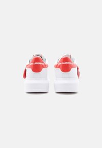 Λευκά αθλητικά παπούτσια με πλατφόρμα, με γυαλιστερές κόκκινες ετικέτες στη φτέρνα, λογότυπο "KARL LAGERFELD" στο πίσω μέρος και κόκκινο γούρι προσαρτημένο στο πλάι.