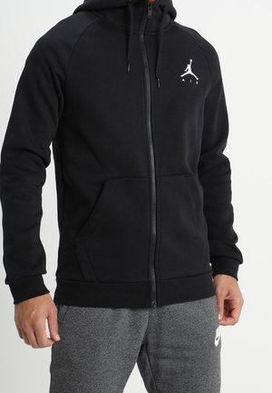 Person iført en sort hættetrøje med lynlås med et hvidt Air Jordan-logo på brystet og grå Nike-sweatpants.