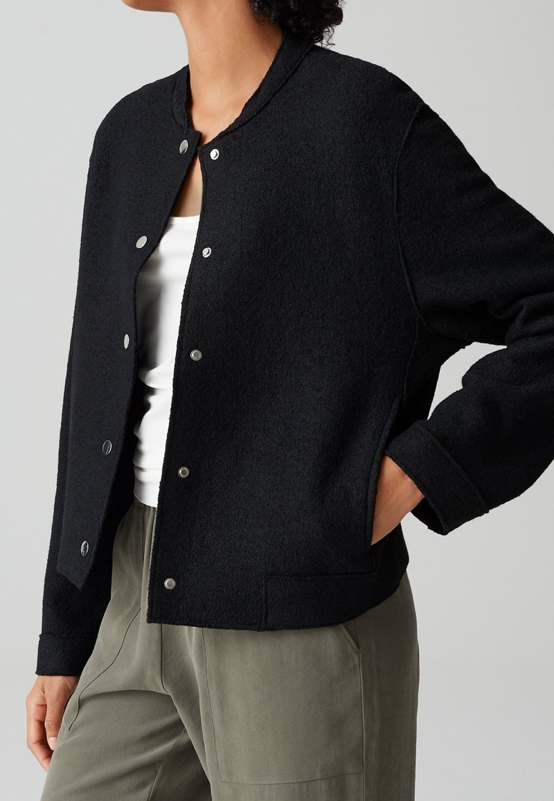 Personne portant une veste noire à boutons-pression sur un top blanc et un pantalon vert olive, une main dans la poche de la veste, sur un fond neutre.