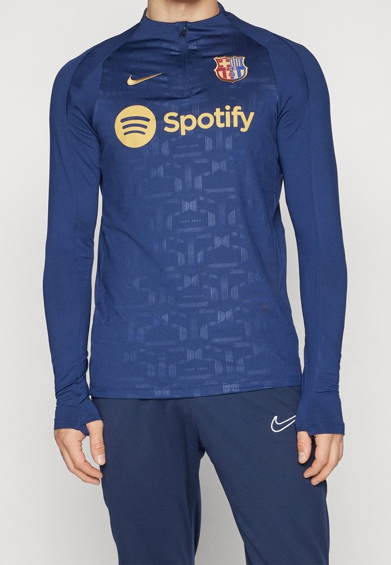 Marineblauwe sporttop met lange mouwen en halve rits, met een gestructureerd patroon, Nike-logo en Spotify-merk. Het materiaal lijkt glad en rekbaar.