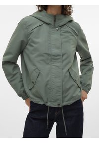Femme portant une veste à capuche vert tamisé avec fermeture éclair à l'avant, boutons-pression, poches à rabat, poignets élastiques et ourlet à cordon sur un jean foncé.