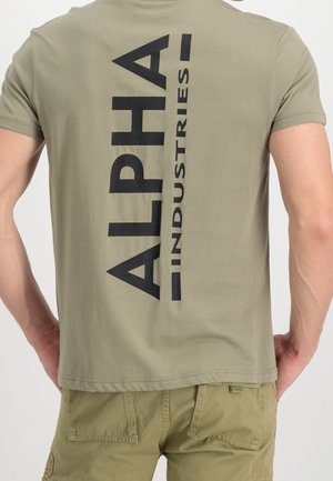 Hombre con camiseta verde oliva con gran texto negro vertical "ALPHA INDUSTRIES" en la espalda y pantalones cortos cargo a juego.
