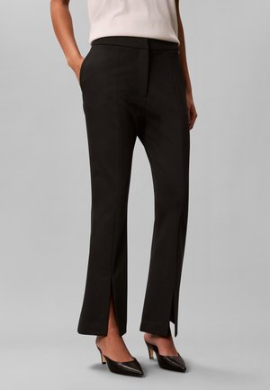 Femme portant un pantalon noir taillé sur mesure avec des fentes à l'avant, associé à des escarpins noirs pointus et un top blanc sans manches.