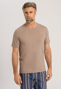 T-shirt marrone a maniche corte con scollo rotondo. Tessuto in cotone morbido, design aderente. Il modello indossa pantaloni a righe in blu, verde e beige.