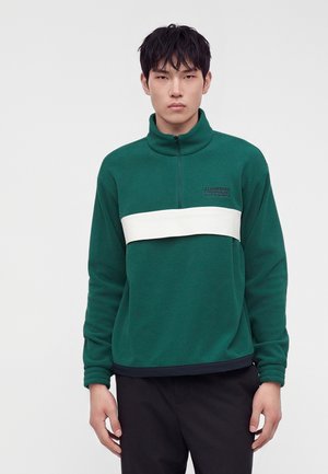 Jeune homme aux cheveux courts et noirs portant un pull polaire vert avec une bande blanche sur la poitrine et un pantalon noir, debout sur un fond blanc.