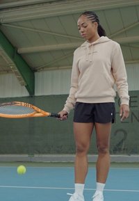 Giocatore di tennis con una felpa beige e pantaloni neri, che tiene una racchetta da tennis arancione, si prepara a colpire una pallina gialla su un campo blu.