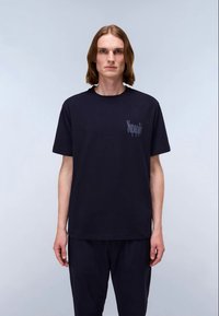 Junge Person mit schulterlangem braunem Haar, die ein schwarzes T-Shirt mit kurzen Ärmeln und einem kleinen Logo auf der Brust trägt, steht vor einem schlichten Hintergrund.
