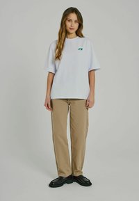 Vit oversized t-shirt med korta ärmar och grön logotyp, parat med beigefärgade vida byxor och svarta skor. Slät tygtextur.