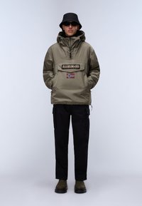 Olijfgroen waterdichte pullover met een groot logopatch en de vlag van Noorwegen, gecombineerd met zwarte cargo-broeken en groene laarzen, gedragen met een zwarte bucket hat.