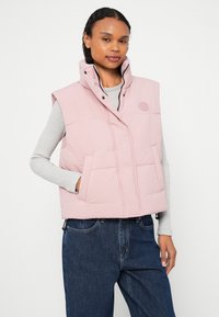 Růžová prošívaná vesta s vysokým límcem, vybavená patentkami, předními kapsami a pod ní žebrovaný svetr. Ležérní střih s hladkou texturou.
