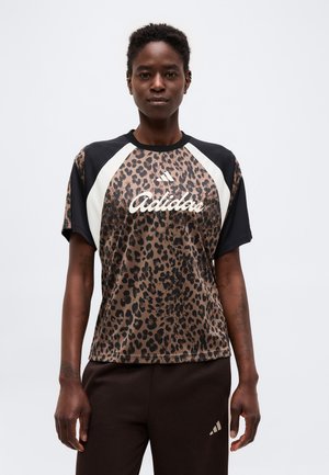 Personne noire portant un t-shirt Adidas à imprimé léopard avec des panneaux noirs et blancs et un pantalon Adidas noir, debout sur fond blanc.