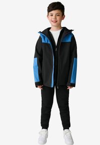 Schwarze Jacke mit blauen Akzenten, mit Reißverschluss vorne, Kapuze und langen Ärmeln. Kombiniert mit schwarzen Hosen und weißen Sneakers. Schlichtes Design.