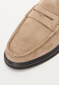 Mocassins en daim beige présentant un bout arrondi, des coutures décoratives et une semelle en caoutchouc noir pour un contraste. Texture lisse, détails de design minimalistes.