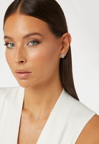 Silberne herzförmige Ohrringe und eine passende Halskette mit einem detaillierten Anhänger, getragen mit einem weißen gerippten Kleidungsstück. Glatte Hautstruktur.