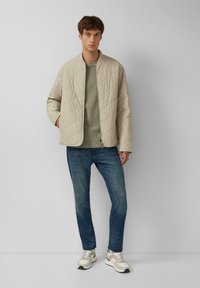 Beige gesteppte Jacke über einem grünen T-Shirt, kombiniert mit blauen Jeans und beigen Sneakers. Einfaches Design mit Rundhalsausschnitt und Reißverschluss.