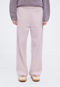 Άτομο που φοράει χαλαρό ανοιχτό ροζ παντελόνι φόρμας και μπεζ αθλητικά παπούτσια, στέκεται μπροστά σε λευκό φόντο, με τα χέρια χαλαρά δίπλα στο σώμα.