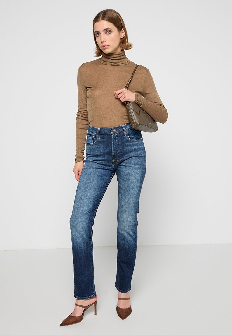 Donna che indossa un maglione a collo alto marrone, jeans blu a vita alta, tacchi marroni e tiene una piccola borsa a spalla marrone, su uno sfondo neutro.