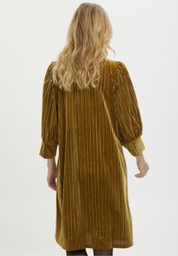 Valkoinen keltainen corduroy-mekko, jossa on pystysuoria raidoituksia, kolmesta neljännestä hihasta ja rento istuvuus. Pituus ulottuu reiteen keskikohtaan.