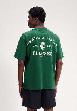 Mężczyzna noszący zieloną koszulkę tenisową klubu Ellesse z napisem "Perugia Italia Dal 1959", widziany od tyłu, w połączeniu z czarnymi szortami.