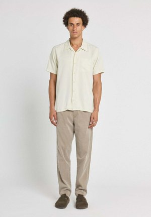 Homme debout, bras détendus, portant une chemise beige à manches courtes, un pantalon marron clair et des chaussures marron foncé sur un fond uni.