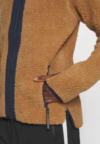 Brązowa polarowa kurtka z granatowymi wykończeniami, z zamkiem błyskawicznym z przodu i kieszeniami po bokach. Wyróżnia się miękką teksturą i przeszyciami.