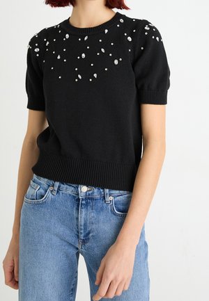 Kobieta ubrana w czarny, dziergany top z krótkim rękawem z rozrzuconymi małymi i dużymi ozdobami z kamieni typu rhinestone w okolicy dekoltu, zestawiony z niebieskimi jeansami.