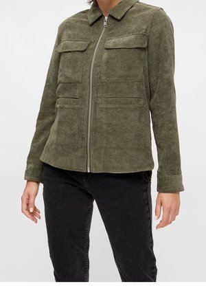 Persona que lleva una chaqueta de pana verde oliva con cuatro bolsillos delanteros y una cremallera central, combinada con pantalones negros.
