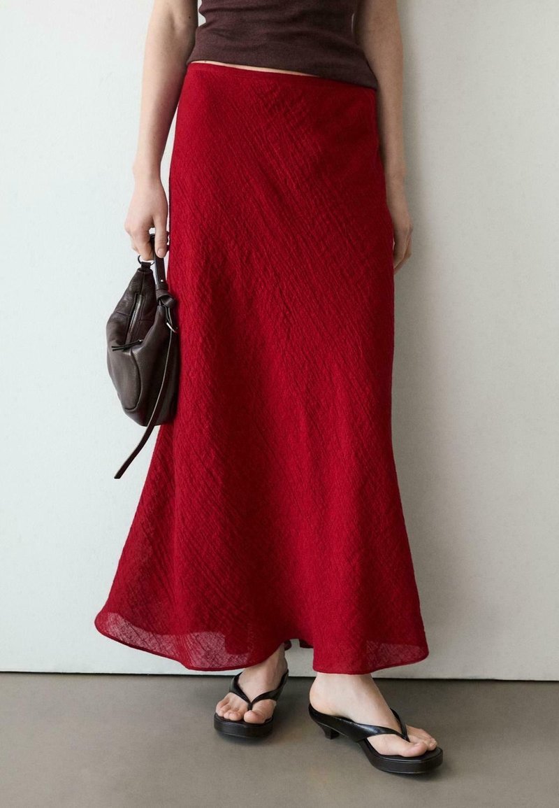 Femme portant une longue jupe rouge texturée, des sandales noires à talons, tenant un petit sac à main noir devant un mur blanc uni.
