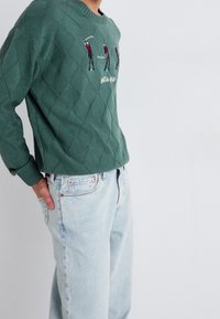 Pull en tricot vert avec motif en losange, orné de figures de golf brodées. Associé à un jean denim bleu clair, affichant une coupe décontractée.