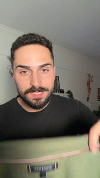 Vīrietis ar bārdu un deguna gredzenu tur zaļu auduma priekšmetu ar melnu etiķeti, smaidot iekštelpās, kura fonā ir sienas dekori un augi.