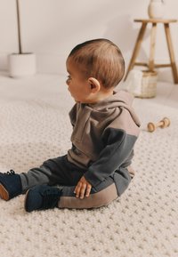 Dwukolorowy strój dla małego dziecka z miękkiego materiału; w kolorach szarym i taupe, z kapturem i dopasowanymi spodniami, oraz ciemnoniebieskimi butami z brązowymi podeszwami, siedzący na teksturowanym dywaniku.