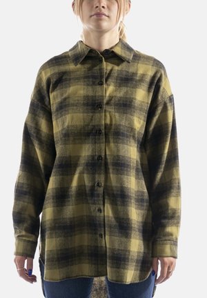 Personne portant une chemise à carreaux verte et noire oversize à boutons avec manches longues et col, associée à un jean bleu.