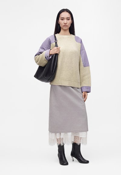 Femme portant un pull beige et lavande, une jupe grise avec un ourlet en dentelle blanche, des bottines noires, et tenant un grand sac à épaule noir.