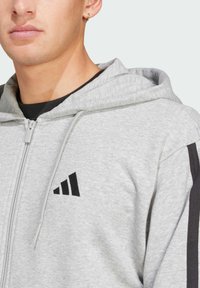 Šedá mikina vyrobená z mäkkého materiálu, s predným zipsom, čiernymi akcentovými pruhmi na rukávoch a čiernym logom na hrudi.