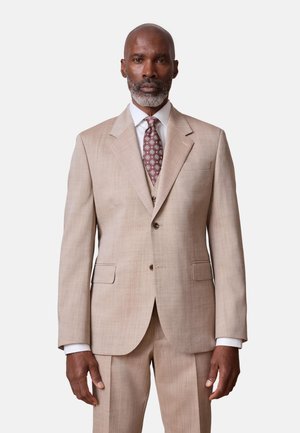 Homme avec barbe grise portant un costume beige, une chemise blanche et une cravate à motifs, debout devant un fond clair uni.