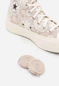 Beżowe trampki Converse wysokie w wzór z białymi akcentami. Zawierają dwie zwinięte beżowe sznurowadła, teksturowany materiał oraz okrągłe oczka.