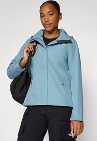 Giacca impermeabile di colore blu chiaro con cappuccio staccabile, tasche con zip e dettagli neri; abbinata a pantaloni cargo neri e a una borsa nera.