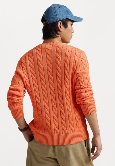 Cable-knit sweater in bright orange, featuring long sleeves, a ribbed waist and cuffs, and a textured finish. Worn over khaki pants.