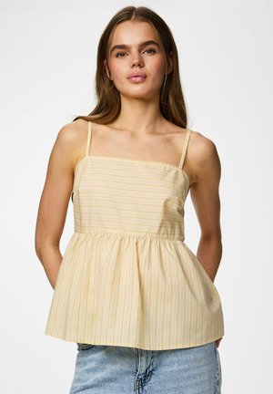 Jeune femme portant un haut peplum sans manches à rayures jaunes et un jean bleu clair, debout devant un fond blanc uni.