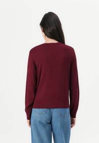 Burgundska izdelava z dolgimi rokavi in krožno ovratnikom, izdelano iz mehke tkanine. Ribljen rob in manšete, v kombinaciji z svetlo modrimi jeans hlačami.