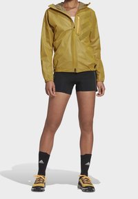 Geel lichtgewicht jack met capuchon, ritssluiting aan de voorkant en zijzak. Gedragen over zwarte shorts en contrasterende zwarte sokken. Getextureerde stof.