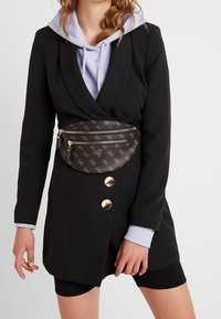 Blazer noir avec des boutons dorés, sweat à capuche violet clair en dessous, et une banane noire à motifs avec des fermetures éclair et un logo.