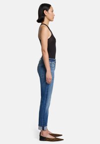 Czarny bezrękawnik, niebieskie dżinsy z podwiniętymi nogawkami i czarne baleriny w panterkę. Modelka pokazana z boku na tle białym.