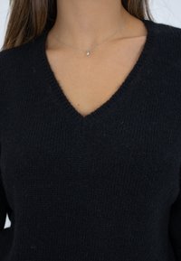 Schwarzer Strickpullover mit V-Ausschnitt und weicher Textur. Hat eine enganliegende Passform. Wird mit einer filigranen silbernen Halskette und einem schlichten Anhänger getragen.